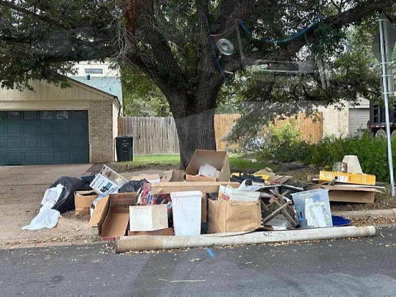A curbside pile of cardboard boxes, plastic bins, trash bags, and broken wood set out under a tree in front of a house with a green garage door