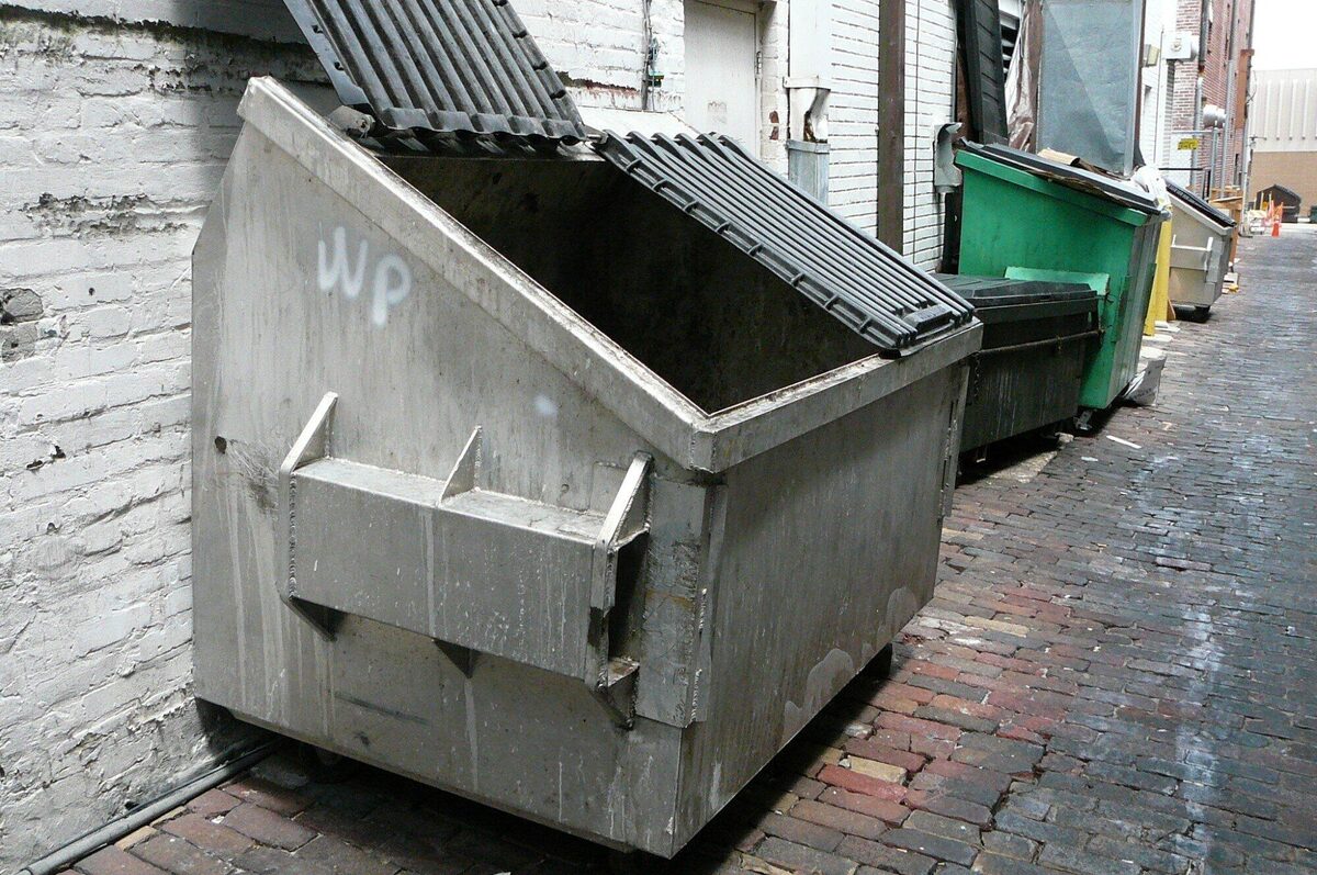 A metal commercial dumpster with its lids open in a brick-paved alley, with more dumpsters lined up behind it