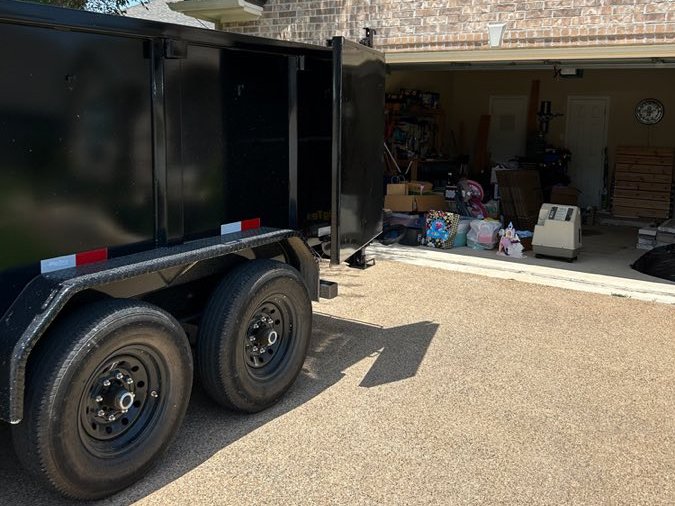 The black BCS Junk dump trailer parked in the driveway of a brick home, beside an open garage filled with boxes, toys, and household items